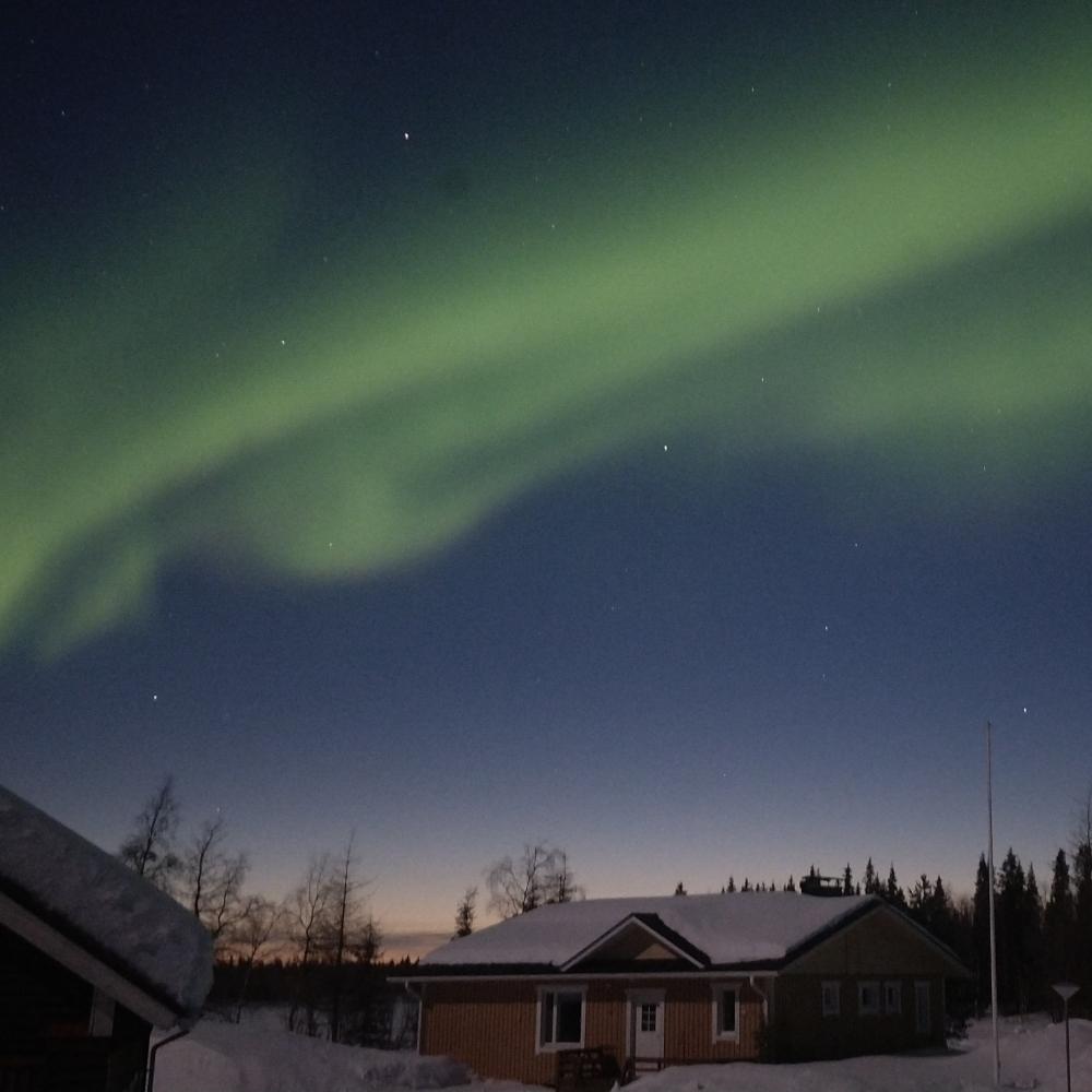 Het noorderlicht boven een huis in Lapland bij schemering, terwijl de oranje gloed van een recente zonsondergang begin april nog steeds aan de horizon te zien is.
