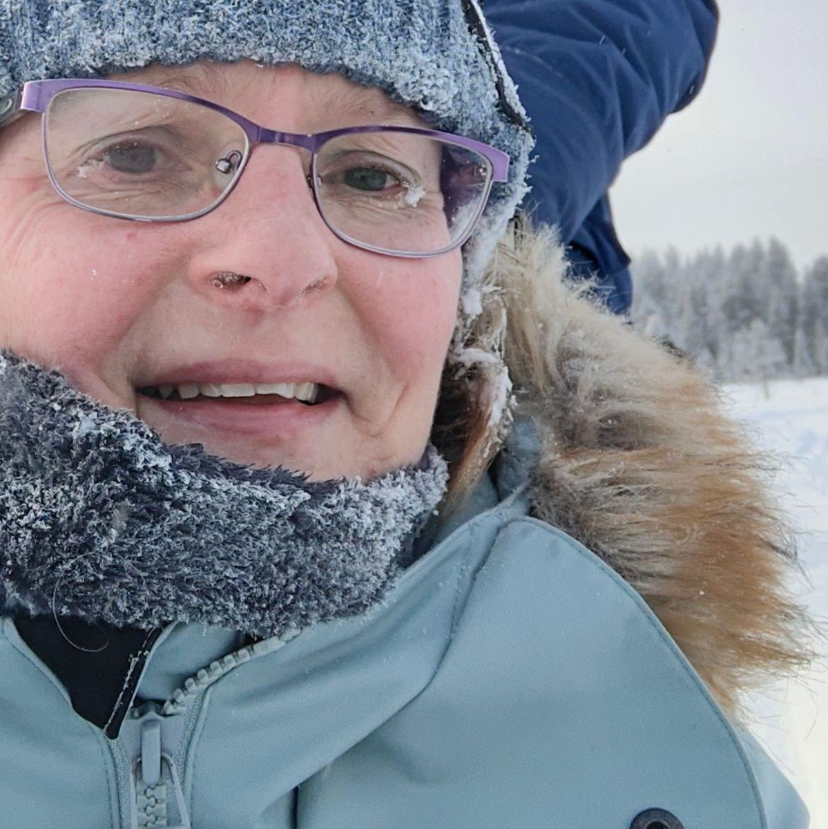 Een close-upportret in Fins Lapland bij extreme kou, waarbij ijskristallen een gebreide muts, sjaal en wimpers bedekken en er zich ijs begint te vormen aan de randen van de bril.