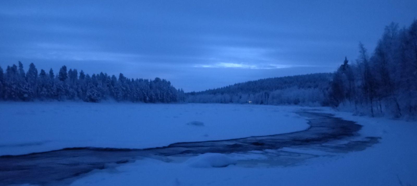 Een bevroren rivier in Lapland tijdens het blauwe uur, half december: een donker, open waterkanaal dat zich een weg baant door de sneeuw en het ijs, met daarachter bossen en heuvels.