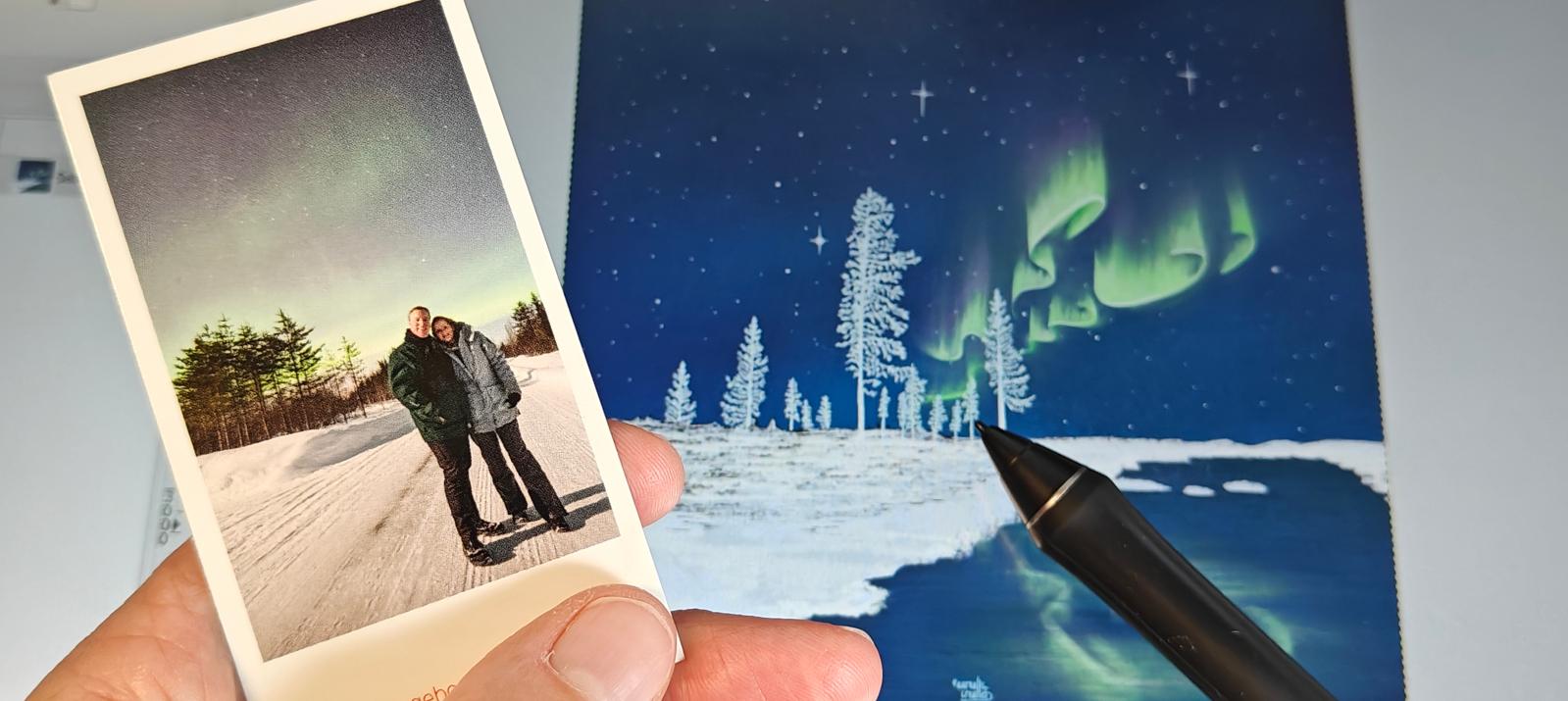 De illustratie ‘Sub-Zero Winter’ op het scherm in GIMP, naast een afgedrukte foto van de kunstenares en haar partner onder het noorderlicht in Fins Lapland, januari 2020. Aurealis Creatief.